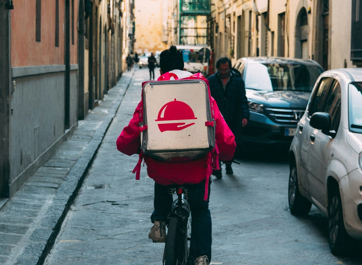 A courier in a red jacket carrying an insulated food backpack through a narrow street