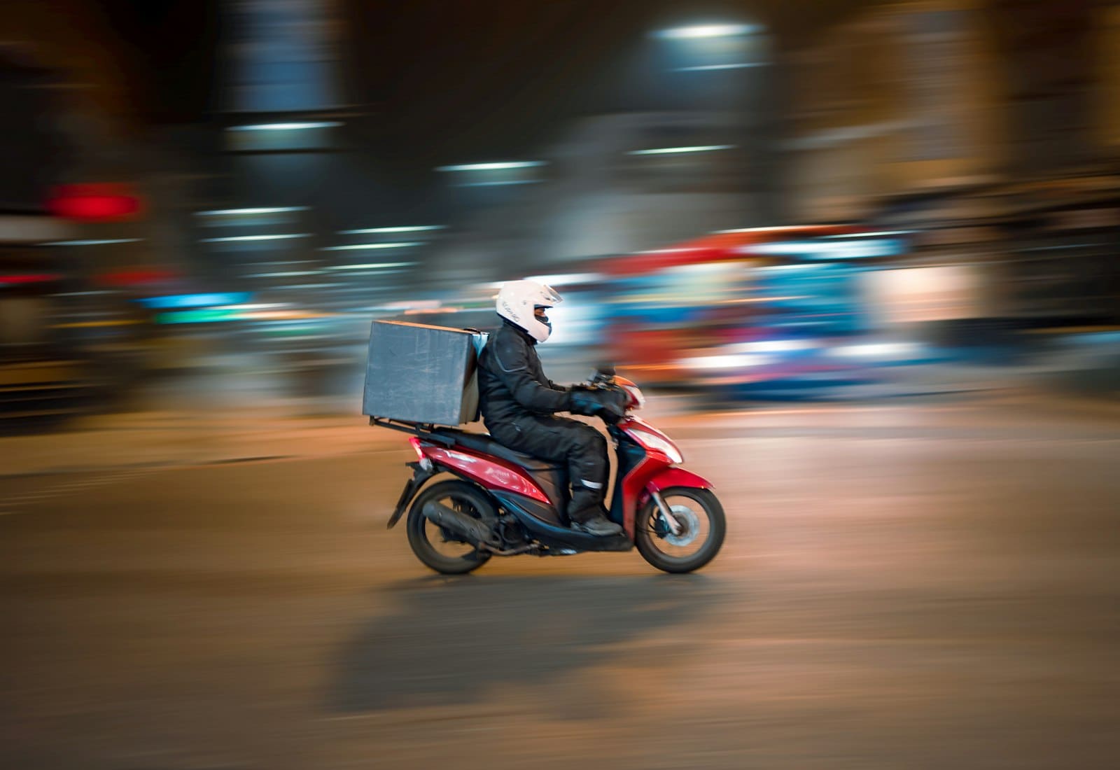 A delivery rider on a red scooter moving through a city at night